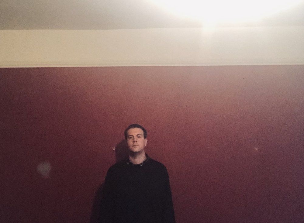 Samuel Nicholson picture, he is standing agains a red wall wearing a black coat. His face looks serious and he is staring at us.